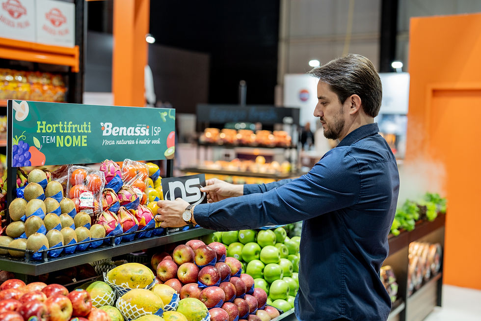 Homem em supermercado repondo frutas; kiwis, maçãs e pitayas são visíveis. Placa "Hortifruti" em destaque. Ambiente iluminado e colorido.