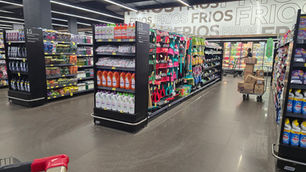 Corredor de supermercado de limpeza, com produtos e vassouras; cliente empurra carrinho entre placas.