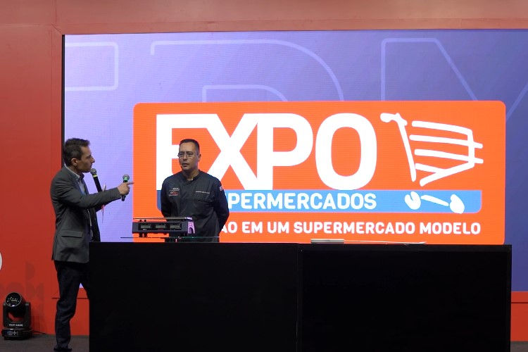 Dois homens num palco, um falando ao microfone, diante de tela com logo EXPO SUPERMERCADOS e texto em fundo laranja e azul.