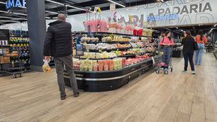 Padaria de supermercado com frutas embaladas e flores; clientes escolhem produtos sob letreiro PADARIA.