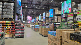Corredor de atacado com pilhas de produtos, telas promocionais da Heineken e placas Cestto; ambiente amplo e iluminado.