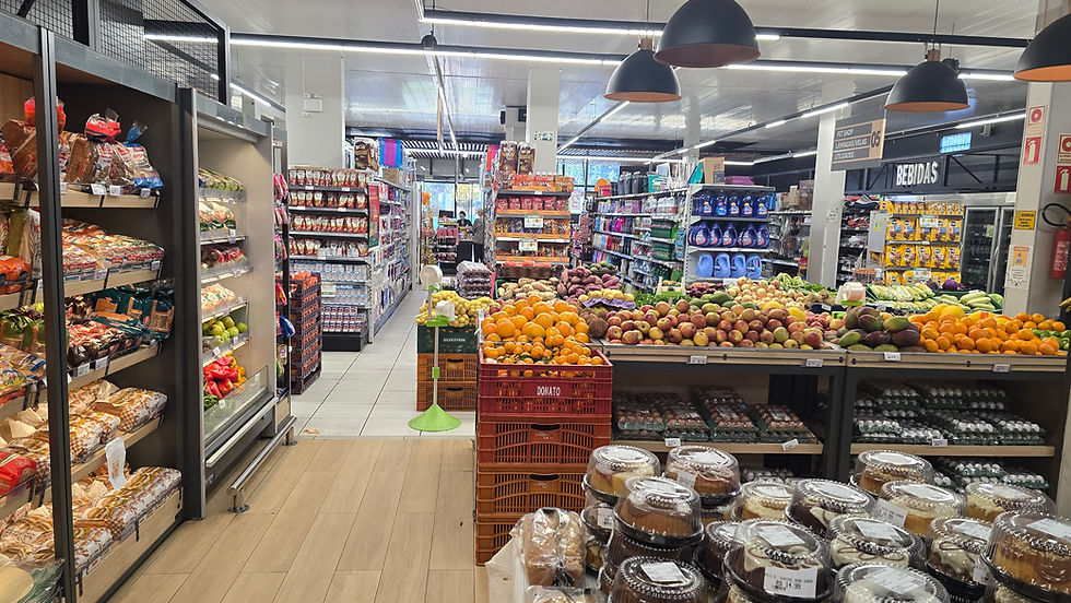 Corredor de supermercado com frutas, verduras e bolos, prateleiras cheias e placa BEBIDAS ao fundo.