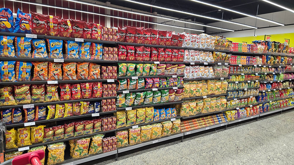 Corredor de supermercado com prateleiras cheias de salgadinhos coloridos, como Doritos, Cheetos e Ruffles, sob luz forte.
