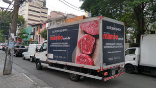 Caminhão de delivery do Mambo com anúncio de carne na rua, em meio ao trânsito e prédios urbanos.
