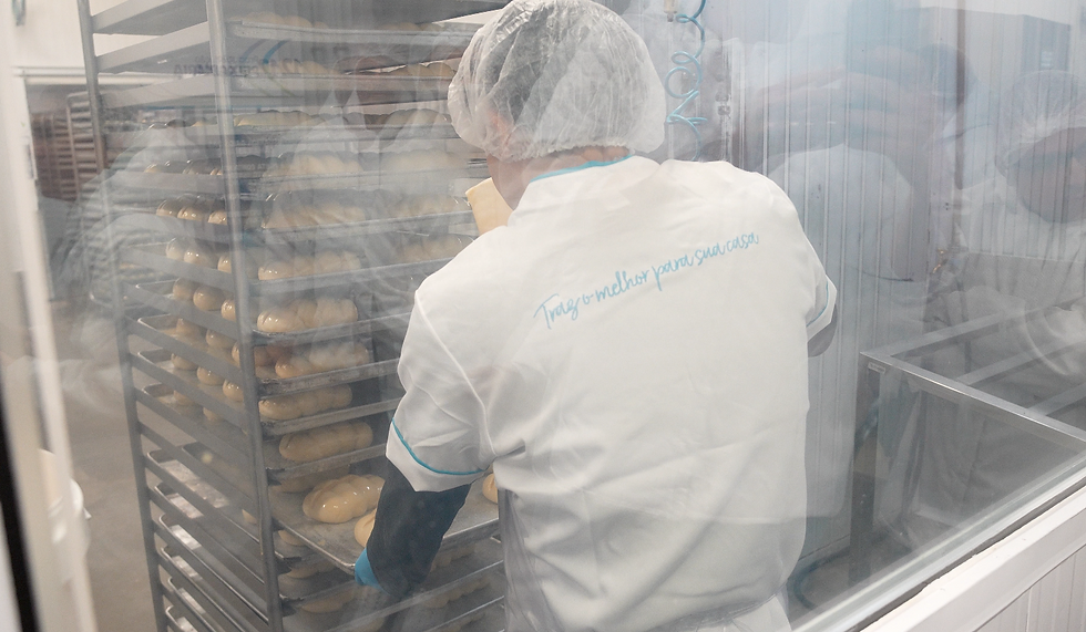 Funcionário de padaria com hairnet e luvas azuis mexe em bandejas de massa num forno industrial; frase azul na camisa.
