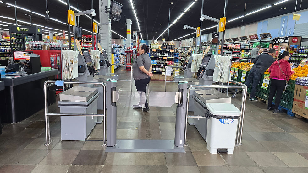 Entrada de supermercado com caixas de autoatendimento; clientes compram frutas e bebidas sob luzes fortes.