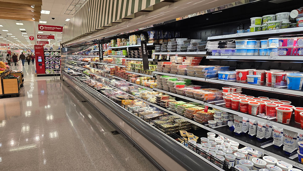 Corredor de supermercado com balcão de laticínios cheio de iogurtes, queijos e creme azedo; clientes ao fundo.
