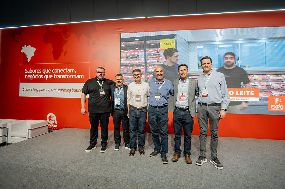 Seis homens posam sorrindo em estande de feira vermelho; ao fundo, texto Sabores que conectam, negócios que transformam.