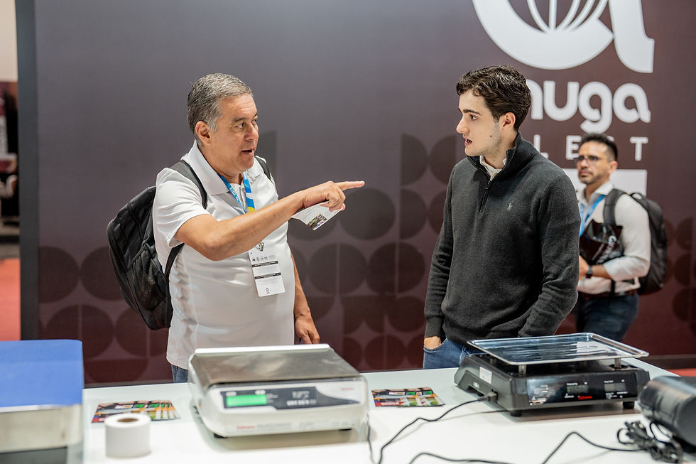 Homem de camiseta branca gesticula para outro de suéter cinza em exposição. Balanças na mesa. Fundo cinza com logo "nuga".