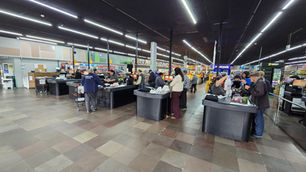 Supermercado movimentado com clientes nas caixas, sob luzes de LED, carrinhos e checkouts pretos.