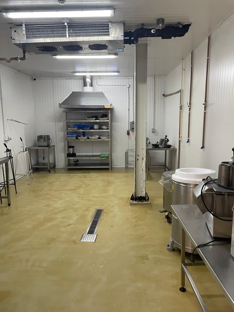 Cozinha industrial vazia com piso bege, prateleiras de metal e equipamentos inoxidáveis. Ambiente limpo e bem iluminado com exaustor no fundo.