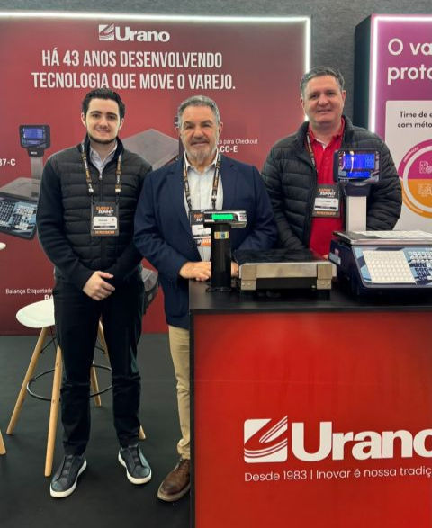 Três homens sorrindo no estande da Urano, entre balanças e equipamentos, com fundo vermelho e textos promocionais.