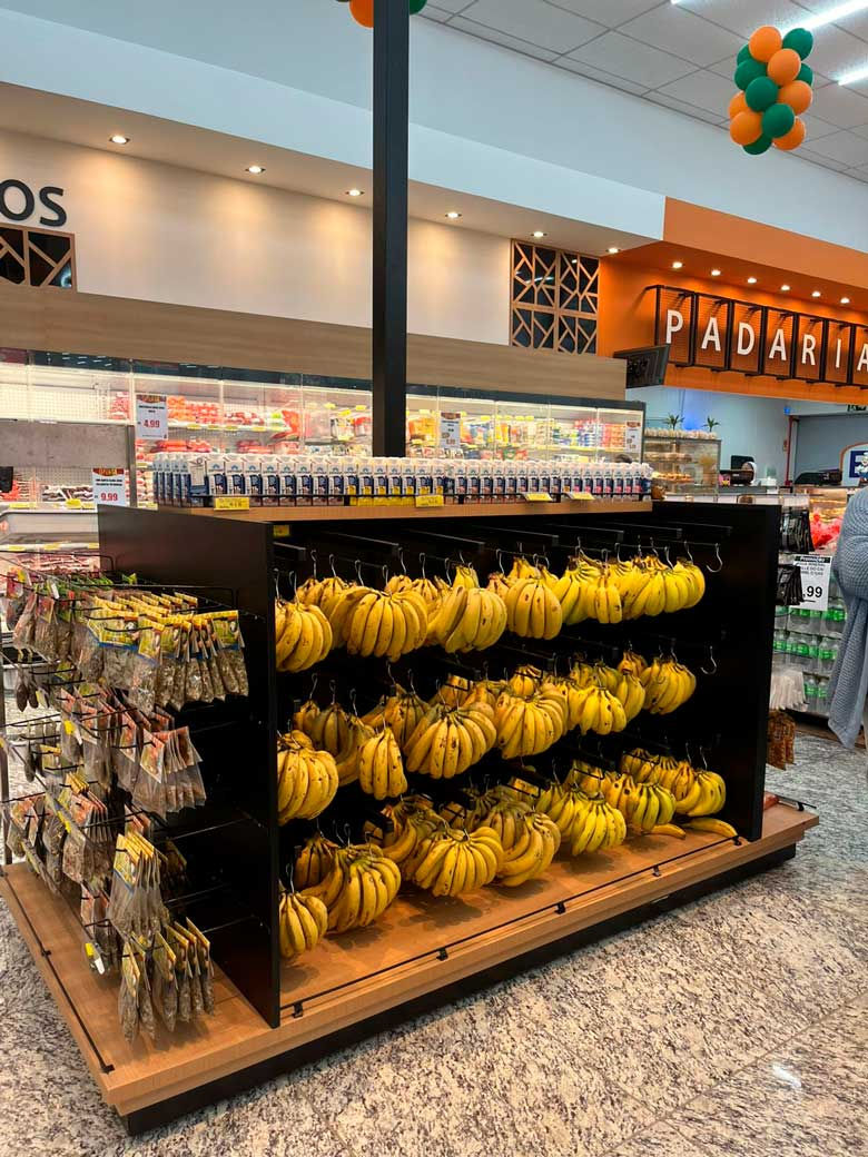 Expositor de bananas em supermercado, com seção PADARIA ao fundo e balões verde e laranja no teto.
