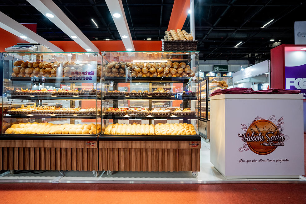 Estande de padaria com vitrines cheias de pães e cestos, em feira coberta; placa Valochi Sousa, ambiente iluminado.