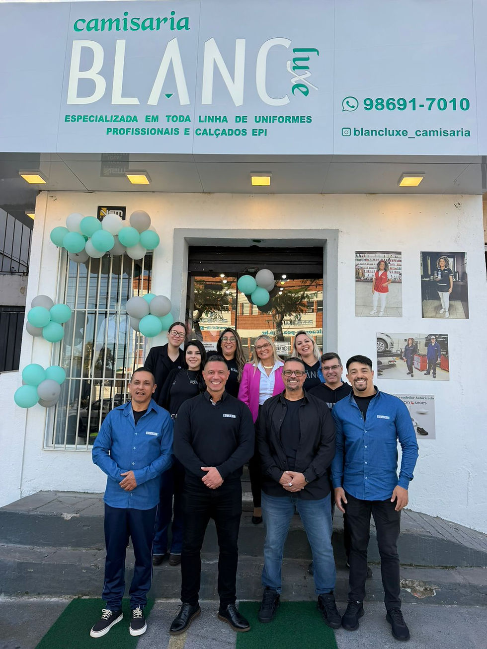 Pessoas sorriem em frente à entrada de uma loja chamada "Camisaria Blanc". Decoração com balões verdes e brancos. Ambiente acolhedor.