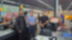 João Dimer, Fabiano Polese e Guilherme Dimer posam no caixa de um supermercado colorido, com frutas, doces e placas AGUARDE! e HORIFRUTI ao fundo.