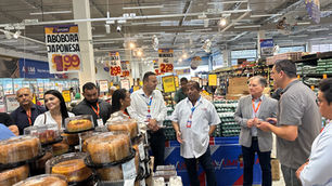 Grupo de pessoas conversa em supermercado AYUMI, entre bolos e promoções de abóbora japonesa, mandioca e cebola.