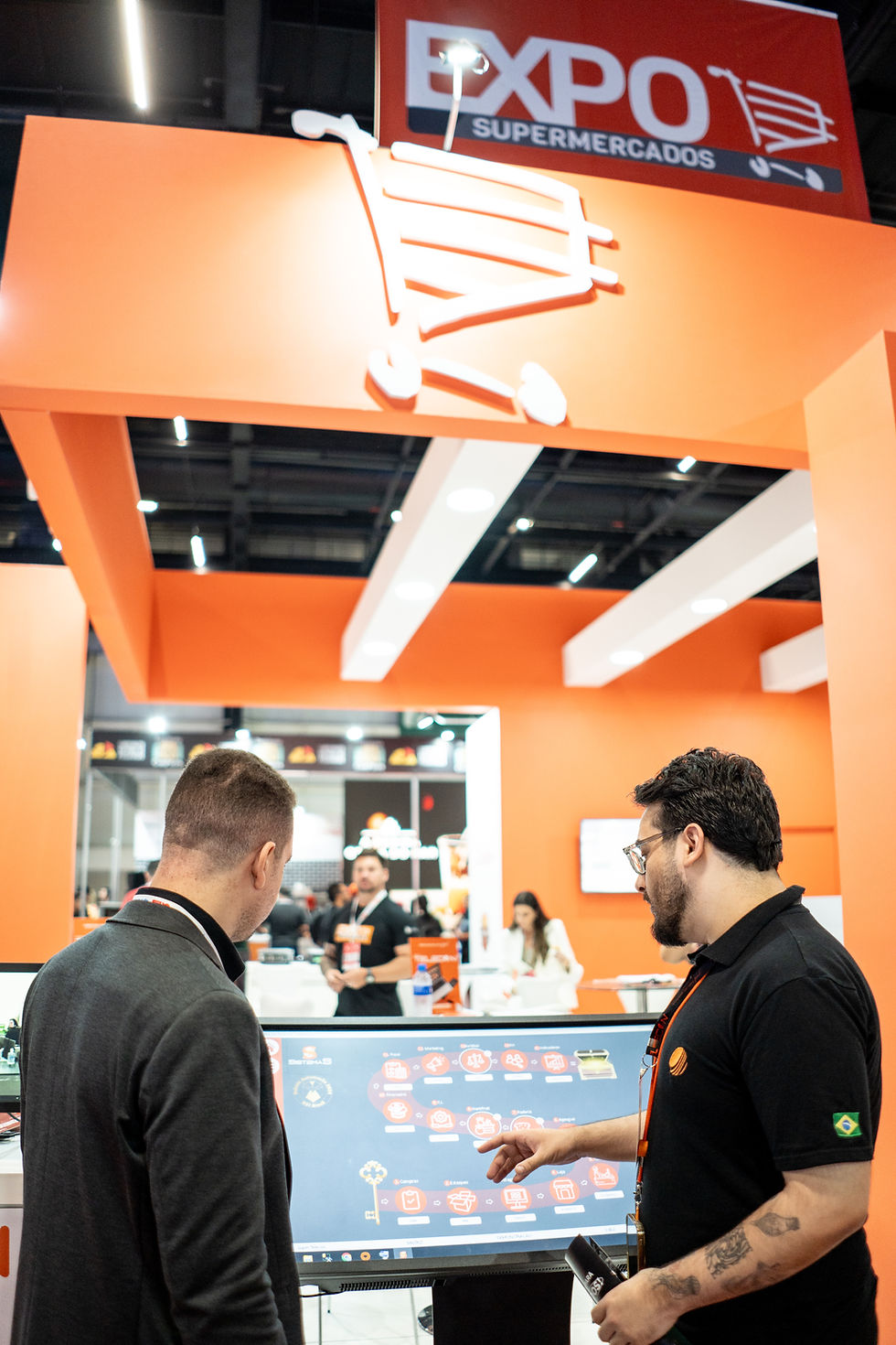 Duas pessoas interagem com tela digital em ambiente de feira. Fundo laranja com logo "EXPO SUPERMERCADOS". Ambiente animado e tecnológico.