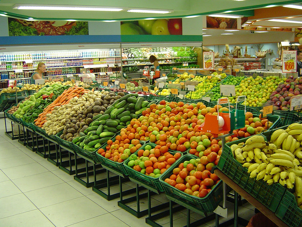 Feira de hortifrúti em supermercado com frutas e legumes coloridos, duas clientes ao fundo e placas de promoção.