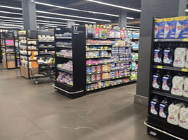 Corredor de loja com prateleiras de cosméticos coloridos; painel com mulher e texto Cuidados Pessoais.