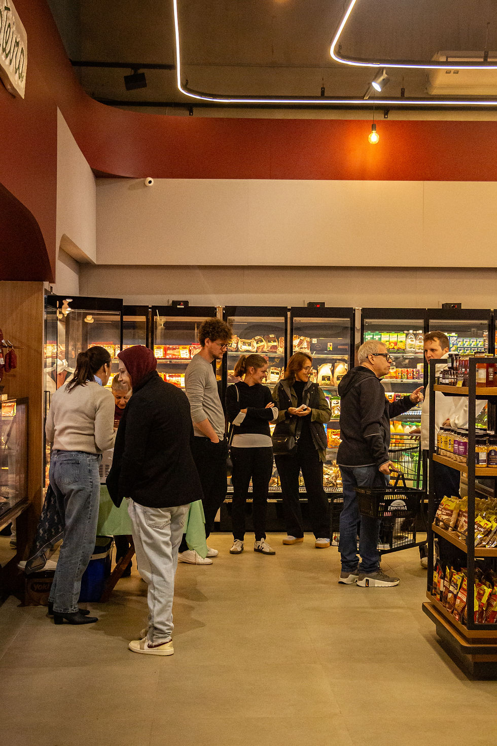Fila de clientes em supermercado iluminado, entre geladeiras e prateleiras, com clima movimentado e casual.