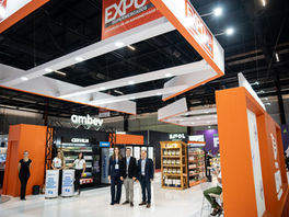 Estande laranja e branco da Ambev em feira, com várias pessoas posando; placas EXPO e CERVEJA, clima de negócios.