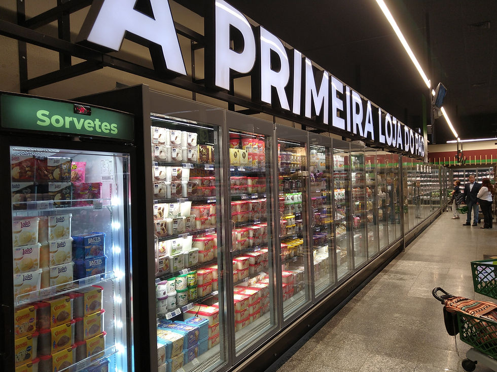 Corredor de freezer de supermercado com sorvetes; placa luminosa A PRIMEIRA LOJA... e clientes ao fundo.
