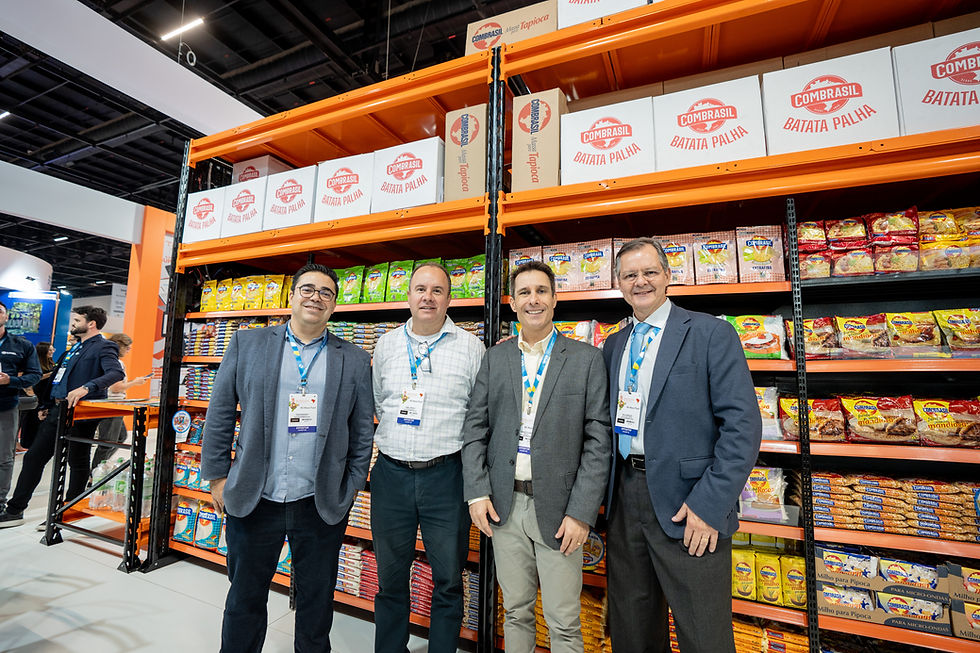 Quatro homens posam sorrindo em frente a prateleiras cheias de produtos alimentícios. Caixas "COMBRASIL" no topo. Ambiente de feira.