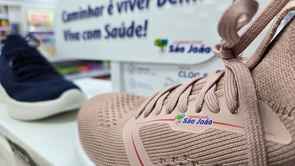 Tênis rosa em destaque com detalhes de Farmácias São João. Ao fundo, placa: "Caminhar é viver bem. Vive com Saúde!" em loja.