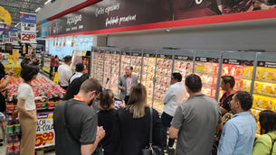 Grupo de adultos em açougue de supermercado, ouvindo Clóvis Polese durante apresentação diante de freezers de carnes e placas de promoção.