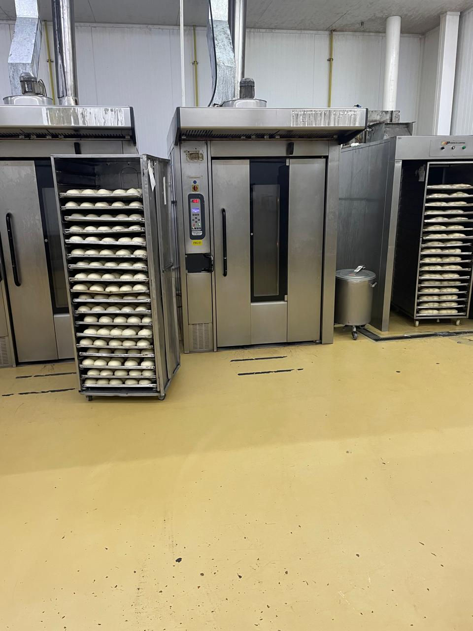 Forno industrial com porta aberta mostra bandejas de pães crus. Piso amarelo claro, paredes brancas, ambiente de padaria moderna.