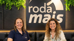 A jornalista Elenise Martins e Luciane, gerente da Makfrio sentadas à mesa, em frente ao letreiro roda mais, em ambiente interno com plantas verdes.