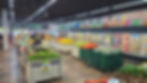 Supermercado com bancas de frutas e legumes coloridos, geladeiras de laticínios ao fundo e clientes circulando.