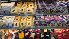 Prateleira de supermercado com vários queijos Vigor, Brie e Gouda em embalagens coloridas e preços visíveis.