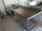 Mesa de inox com pia e prateleiras em uma cozinha. Piso cerâmico, caixa de papelão e mosaico ao fundo. Ambiente organizado e claro.
