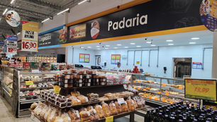 Padaria de supermercado com pães, doces e bebidas expostos; funcionários ao fundo e placas de oferta em destaque.