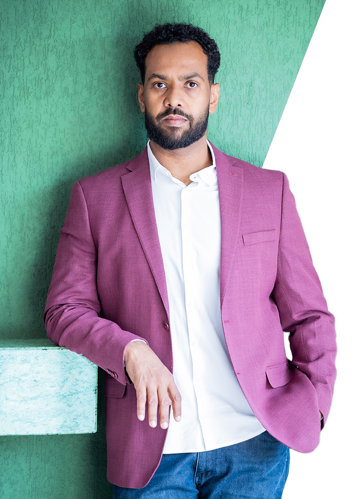 Bruno Cruz, vestindo blazer roxo e camisa branca, encostado em parede verde. Expressão séria. Fundo com textura suave.