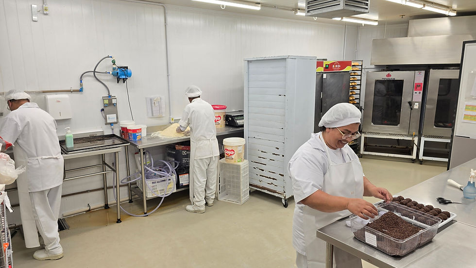 Três pessoas em uma padaria. Duas preparam massa; uma molda doces com chocolate. Ambiente claro, uniforme branco, pote “Soya”. Atmosfera concentrada.