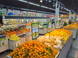 Hortifruti de supermercado com laranjas, melões e uvas em bancas, placas de oferta em português e luzes no teto.