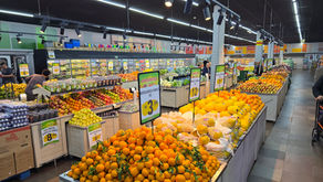 Hortifruti de supermercado com laranjas, melões e uvas em bancas, placas de oferta em português e luzes no teto.
