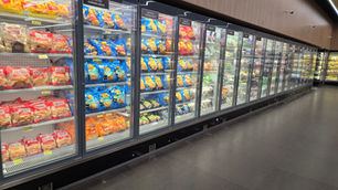 Corredor de supermercado com freezers cheios de batatas, legumes e pães congelados, embalagens coloridas e placas em português.