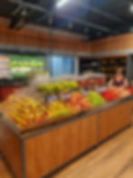 Banca de frutas em supermercado, com uvas, maçãs e bananas; mulher atende ao fundo, em ambiente moderno e iluminado, com névoa fria