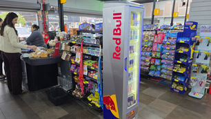 Interior de loja de conveniência com duas mulheres no caixa, geladeira Red Bull e expositores de doces Haribo, clima movimentado.