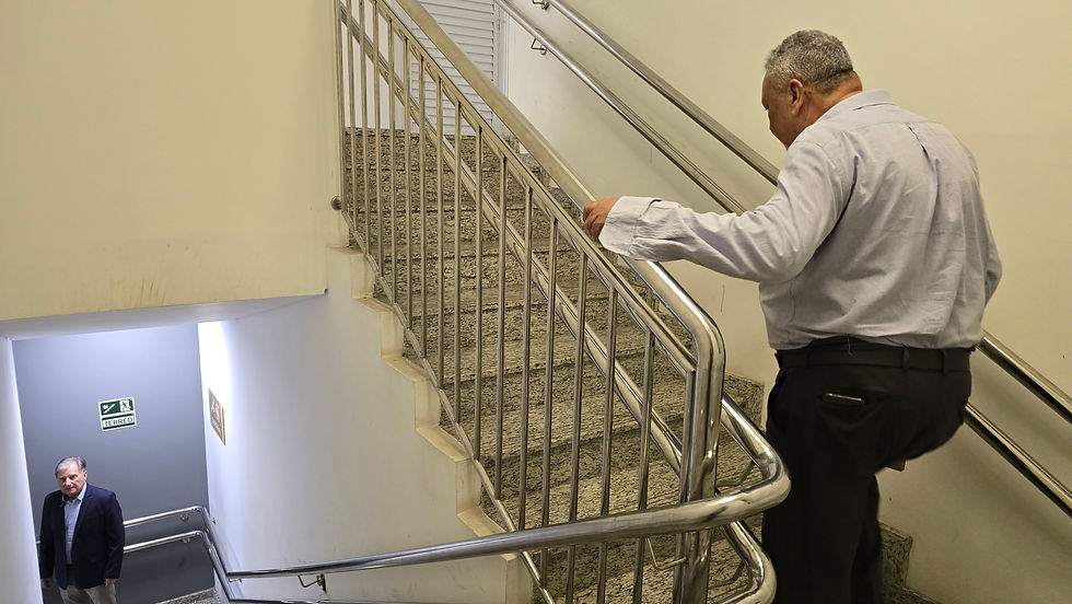Homem descendo escada cinza com corrimão de metal. Outro homem sobe, vestindo camisa clara. Placa de saída de emergência ao fundo.