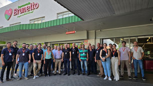 Visita Técnica com grupo de pessoas posando na entrada do supermercado Brunetto, com placas Banco24Horas e fachada branca e verde.