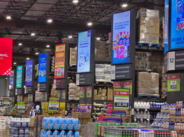 Corredor de supermercado com pilhas de produtos de limpeza, telas promocionais coloridas e placas de preços.