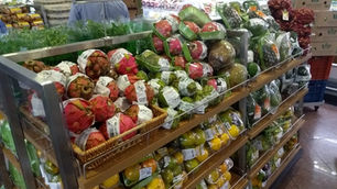 Banca de hortifruti em supermercado, com frutas e legumes embalados, ervas, placas de orgânicos e ovos ao fundo.