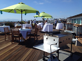 A photo of a rooftop deck with Green Umbrellas, white table cloth covered tables, and the GUAC Taco catering grill