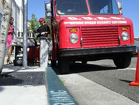 Photo of the GUAC Taco truck parked up against a curb, with an employee serving a Taco to a customer.