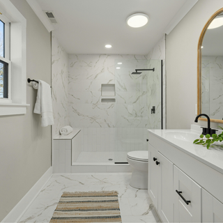 Bright modern white bathroom with marble-tile shower, black fixtures, wood-framed mirror, vanity, plant, and striped rug.